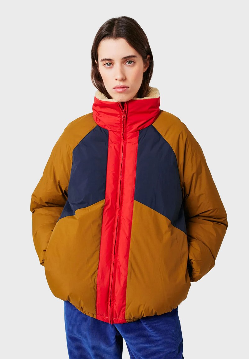Femme portant une veste matelassée en coloris blocs avec des sections rouge, marine et marron, se tenant les mains dans les poches devant un fond uni.