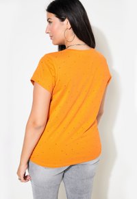 Orange kortärmad t-shirt med rund halsringning och svarta detaljer, i kombination med grå tajta byxor. Bakifrån visar den en slät tygtextur.