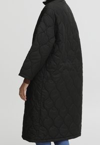 Abrigo negro acolchado de diseño largo y oversize, con un cuello alto y patrón texturizado, mostrado de perfil con mangas en contraste.