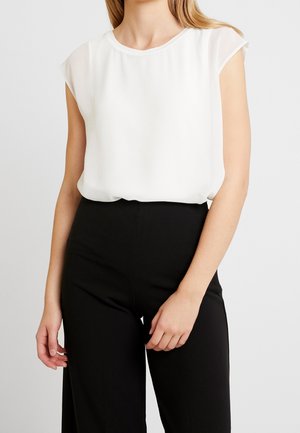 Žena v bílé haleně bez rukávů zastrčené do černých kalhot s vysokým pasem na jednobarevném pozadí.