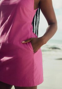 Rosa bomullslös klänning utan ärmar med sidofickor, lös passform och en mjuk textur, visad mot en strandbakgrund.