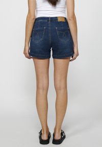 Temno denim kratke hlače z visokim pasom, zavihanimi robovi in dekorativnim šivanjem na zadnjih žepih. Imajo usnjen oznako blagovne znamke na pasu.