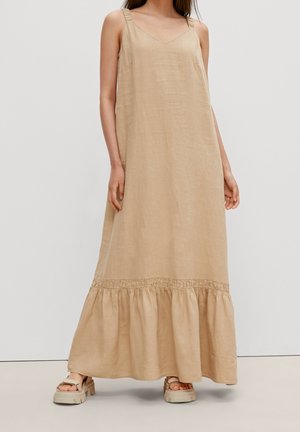 Woman wearing a beige sleeveless maxi dress with ruffled hem and beige platform sandals, standing against a plain light background.