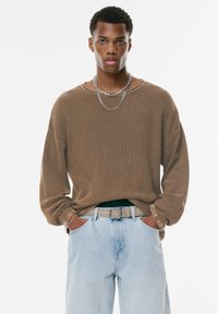Brauner, strukturierter, langärmliger Pullover mit lockerer Passform, kombiniert mit hellblauen Jeans und einem grauen Gürtel. Zwei übereinander liegende Halsketten sind sichtbar.