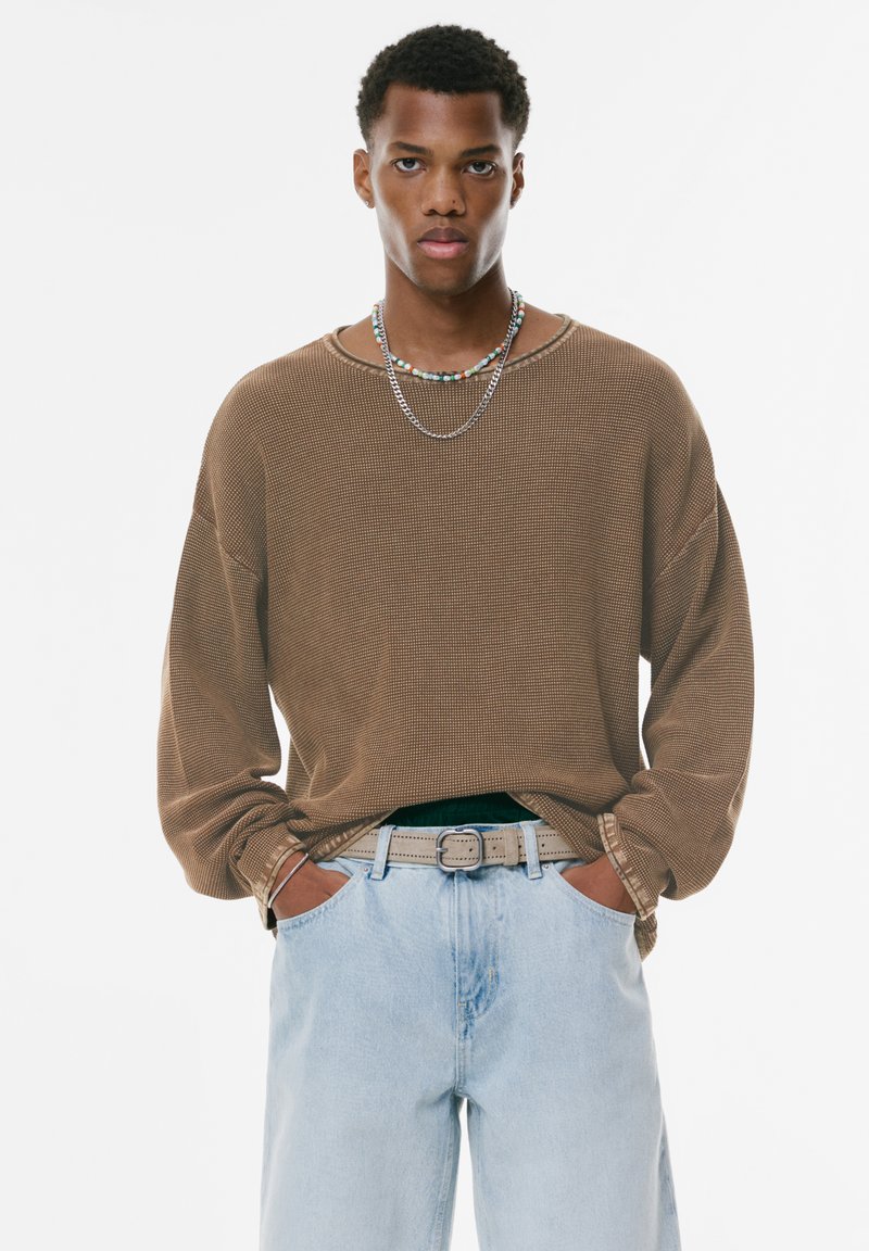 Brauner, strukturierter, langärmliger Pullover mit lockerer Passform, kombiniert mit hellblauen Jeans und einem grauen Gürtel. Zwei übereinander liegende Halsketten sind sichtbar.