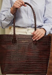 Sac fourre-tout marron foncé avec une texture crocodile, doté de deux longues poignées et d'un logo doré à l'avant, pr�ésenté sur un fond clair.