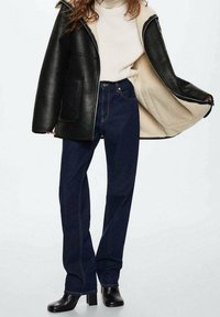 Giacca nera in similpelle con fodera in shearling, dolcevita crema, jeans dritti blu scuro e stivali neri con tacco alla caviglia.