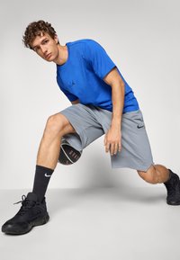 Tricou albastru cu logo-ul Jordan negru, pantaloni scurți atletici gri, încălțăminte de baschet neagră, ținând o minge de baschet cu suprafață texturată, fundal de studio.