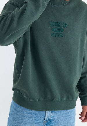 Osoba w ciemnozielonej bluzie z napisem „Brooklyn 1993 New York”, w połączeniu z jasnoniebieskimi jeansami, ręka blisko twarzy.