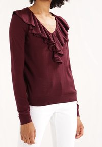 Suéter burdeos con escote en V, cuello con volantes y mangas largas, elaborado en una tela suave, combinado con pantalones blancos.