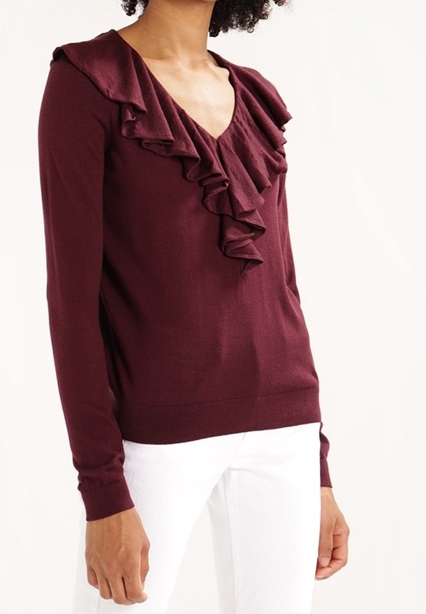 Suéter burdeos con escote en V, cuello con volantes y mangas largas, elaborado en una tela suave, combinado con pantalones blancos.