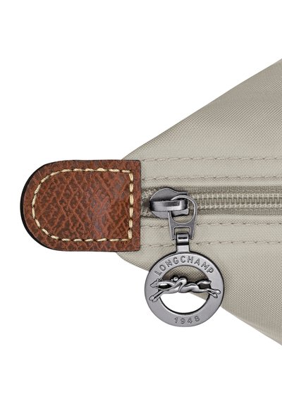 Beige fabric bag corner with brown leather trim and a silver Longchamp zipper pull featuring a horse and rider logo, dated 1948.