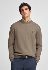 Maglione beige a girocollo realizzato in morbido tessuto a maglia, con un piccolo logo blu marine sul petto, abbinato a una camicia con colletto e pantaloni scuri.