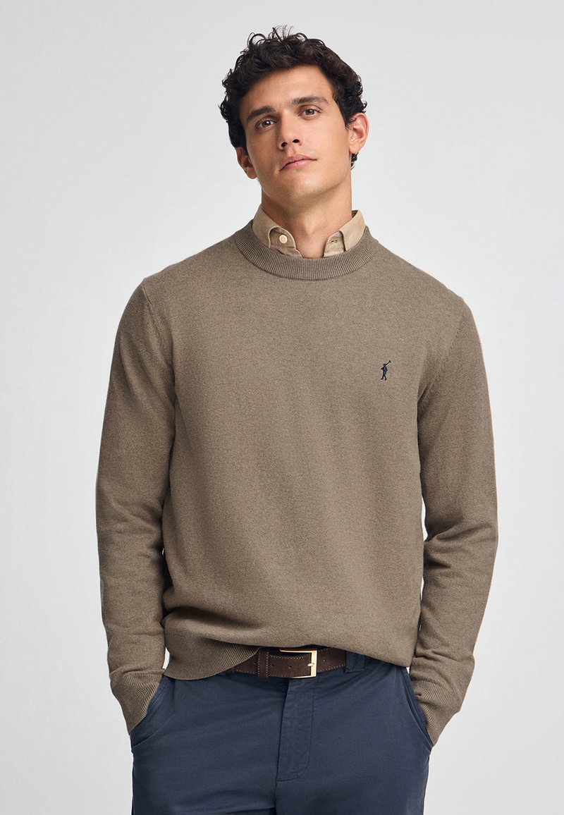 Maglione beige a girocollo realizzato in morbido tessuto a maglia, con un piccolo logo blu marine sul petto, abbinato a una camicia con colletto e pantaloni scuri.