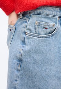 Χέρι σε τσέπη ανοιχτό μπλε τζιν παντελονιού με λογότυπο Tommy Hilfiger, συνδυασμένο με κόκκινο πλεκτό πουλόβερ.