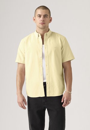 Man die een lichtgele kortebare blouse draagt over een wit tanktopje met een zwarte broek, staand tegen een effen achtergrond.