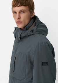 Grijze geïsoleerde jas met een capuchon, voorzien van een ritszak, verstelbaar trekkoord en logo-embleem op de mouw. Gladde textuur.