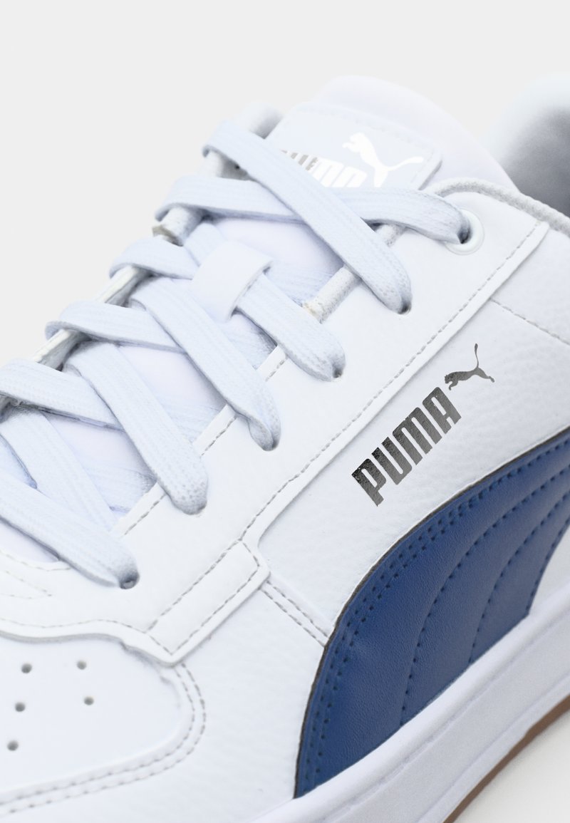 Sneaker en cuir blanc avec des accents bleus, panneau latéral texturé, lacets gris et logo argenté sur le côté. Design minimaliste.