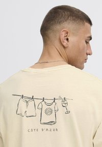 Lysegul t-shirt med et grafisk motiv af svømmebukser, en t-shirt og en drink hængende på en tørresnor med "CÔTE D'AZUR" tekst nedenfor.