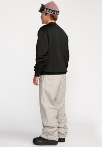 Sweatshirt vert foncé, pantalon gris clair ample et bottes noires. Un bonnet rose clair avec un bandeau en logo noir et des lunettes sur le front.