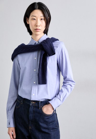 Young person with short black hair wearing light blue buttoned shirt, dark blue jeans, and a navy cable knit sweater draped over shoulders.