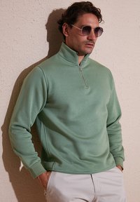 Green sweatshirt with a zip collar, soft texture, long sleeves, and a relaxed fit, paired with light-colored pants and sunglasses.