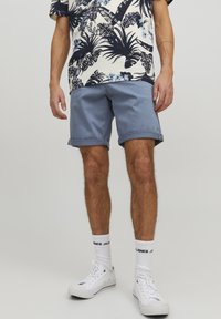 Pantaloni corti azzurri chiari con orli risvoltati, indossati con una camicia a fantasia crema e blu navy e sneakers bianche. Il modello si trova davanti a uno sfondo neutro.