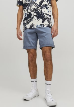 Pantalones cortos azul claro con dobladillos enrollados, combinados con una camisa de patrón crema y azul marino y zapatillas blancas. El modelo está de pie en un fondo neutro.