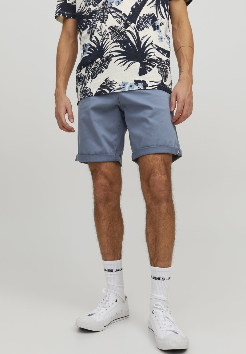 Pantaloni corti azzurri chiari con orli risvoltati, indossati con una camicia a fantasia crema e blu navy e sneakers bianche. Il modello si trova davanti a uno sfondo neutro.