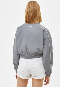 Šedá zkrácená bunda s hladkou texturou, elastickým lemem a bílými bočními pruhy. Nositá přes bílé šortky, pohled zezadu.