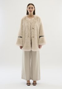 Faux-Fur-Mantel in hellbeige mit flauschiger Textur, der einen Schalkragen und weite Ärmel hat. Schwarze Akzente entlang der Bündchen.