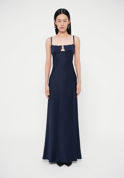 Vrouw staand, gekleed in een lange marineblauwe jurk met dunne bandjes en een uitsnijding onder de gestructureerde borstcups, gecombineerd met hangende oorbellen.