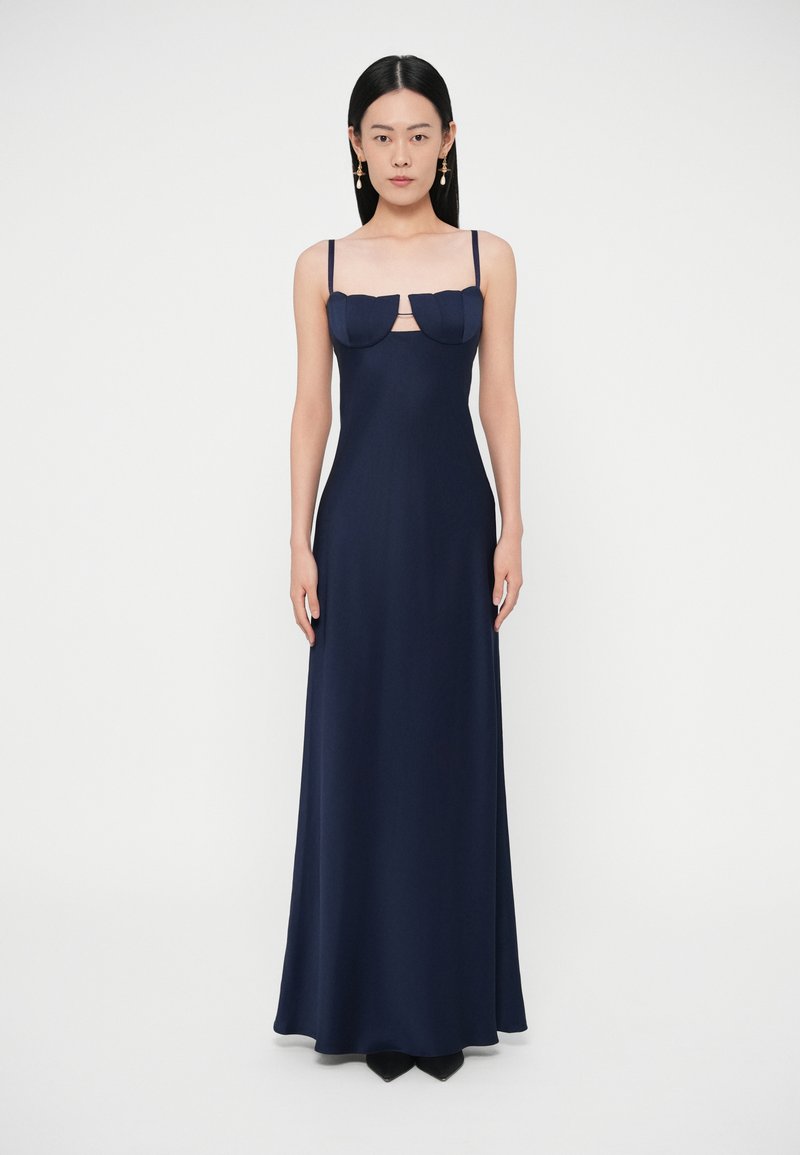 Sieviete stāv, valkājot garu tumši zilu kleitu ar plānām lencēm un izgriezuma detaļu zem strukturētām krūšu kausiņām, valkājot kritiena auskarus.