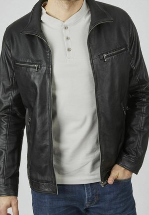 Homme portant une veste en cuir noire dézippée par-dessus un t-shirt henley blanc et un jean bleu, une main dans la poche, torse visible.