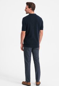 Tummansininen lyhythihainen polo-paita, jossa on teksturoitu neule, yhdistettynä tummiin housuihin ja ruskeisiin kenkiin, nähtynä takaa.