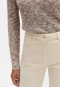 Gehaakte trui in bruine en beige tinten, gecombineerd met lichtbeige broeken met een knoopsluiting en zakken aan de zijkant.