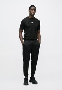 Melns sportisks apģērbs, tajā ir īsām piedurknēm pieguļošs krekls un šaurie bikses. Krekla dizainā ievērojams logotips; apavi ir melni ar teksturētiem akcentiem.