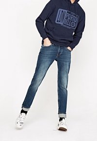 Osoba oblečená v námořnické mikině s grafickým textem, s krátkými modrými džíny, černými ponožkami a bílými teniskami s černými vzory, s rukama v kapsách.