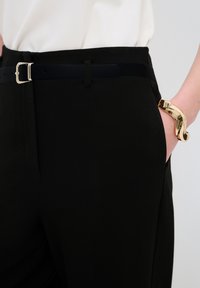 Schwarze Hose mit glatter Textur, ausgestattet mit einem silbernen Schnallen-Gürtel. Ein glänzendes goldenes Armreif ist teilweise an der Hand sichtbar.
