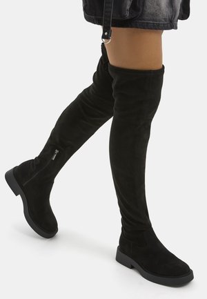Mujer que lleva botas negras de ante por encima de la rodilla con tacones bajos y gruesos y cremalleras laterales, combinadas con una falda negra.