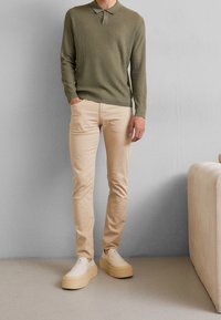 Olive green knit long-sleeve polo shirt, beige slim-fit chinos, and beige platform slip-on shoes, standing against a light gray background.