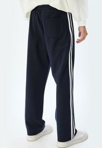 Pantalones de chándal en navy con cintura ajustable, que presentan franjas laterales blancas y un bolsillo trasero. Tejido suave, corte holgado y diseño de pierna recta.