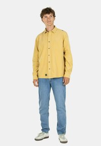 Camisa de botonadura amarilla de pana con botones negros, combinada con jeans azul claro y zapatillas grises. Presenta una etiqueta de logo en el lado izquierdo.