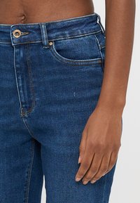 Σκούρο μπλε τζιν με στενή εφαρμογή, που διαθέτει χρυσό κουμπί για κλείσιμο, πέντε τσέπες και διακριτικές ραφές στις ραφές.