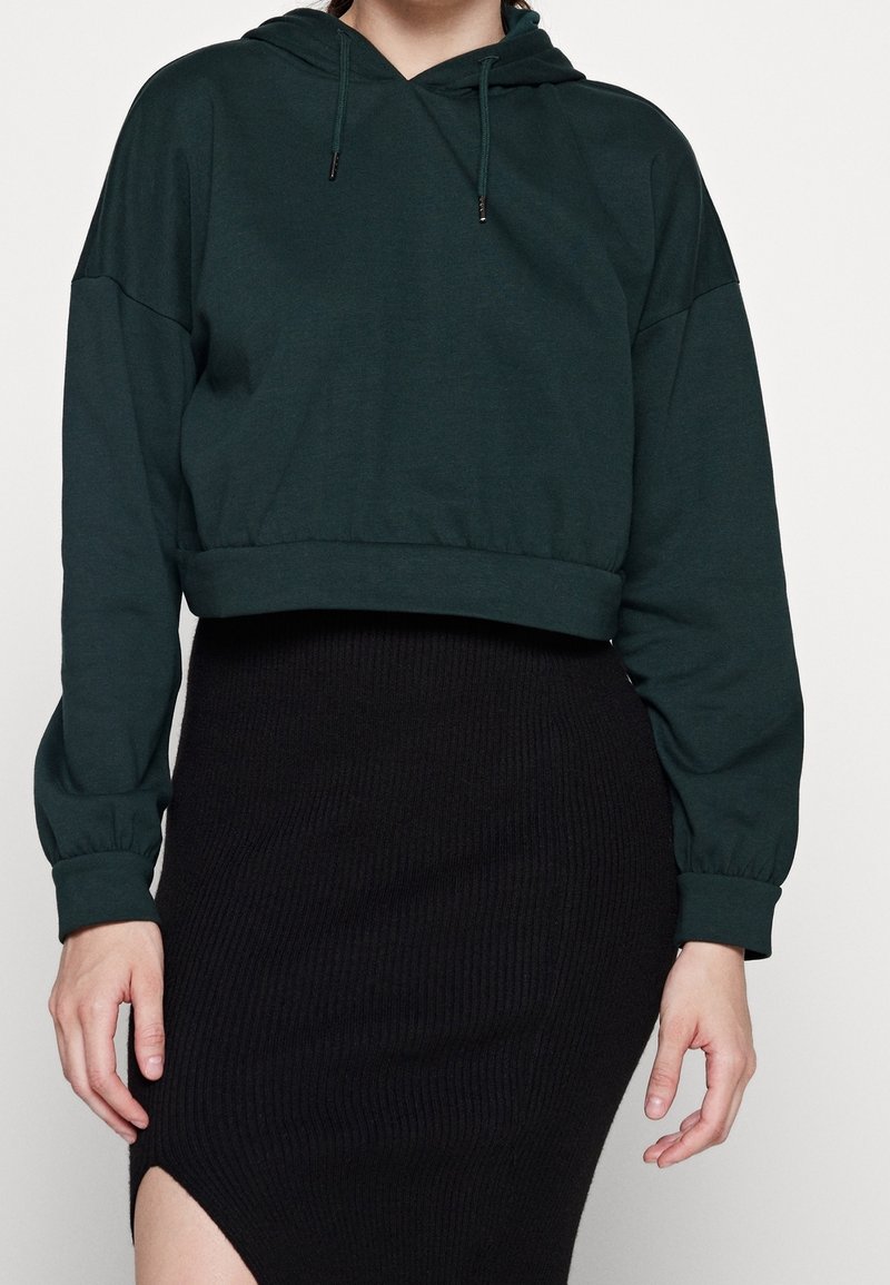Sudadera recortada de color verde oscuro con capucha estructurada, mangas largas y puños elásticos, combinada con una falda negra acanalada con una apertura lateral.