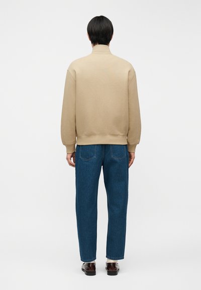 Άνδρας με μαύρα μαλλιά, φορώντας μπεζ φούτερ, μπλε τζιν, άσπρες κάλτσες και σκούρα καφέ παπούτσια, στέκεται με την πλάτη σε ένα απλό λευκό φόντο.