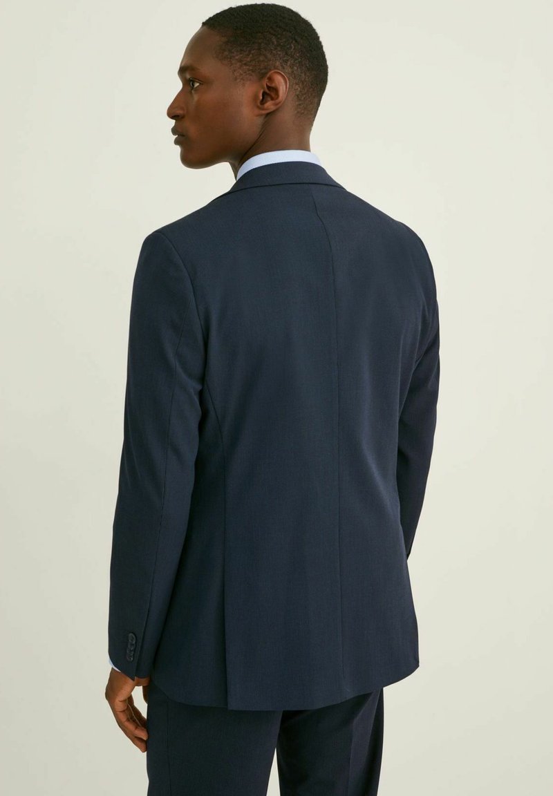 Man som bär en mörk marinblå kostymjacka och byxor, stojande i profil vänd mot vänster mot en enfärgad ljus bakgrund.