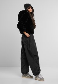 Veste à capuche en fausse fourrure noire, pantalon large noir et baskets grises avec des rayures blanches. Le mannequin porte un bonnet noir et des lunettes de soleil.