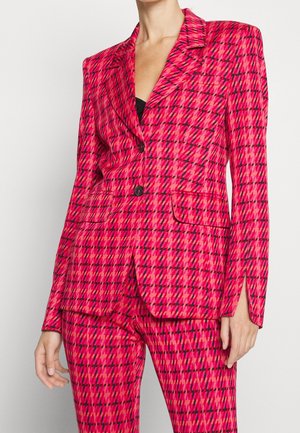 Mujer con blazer a cuadros rosa fuerte y rojo y pantalones a juego con detalles de botones negros y top negro debajo.