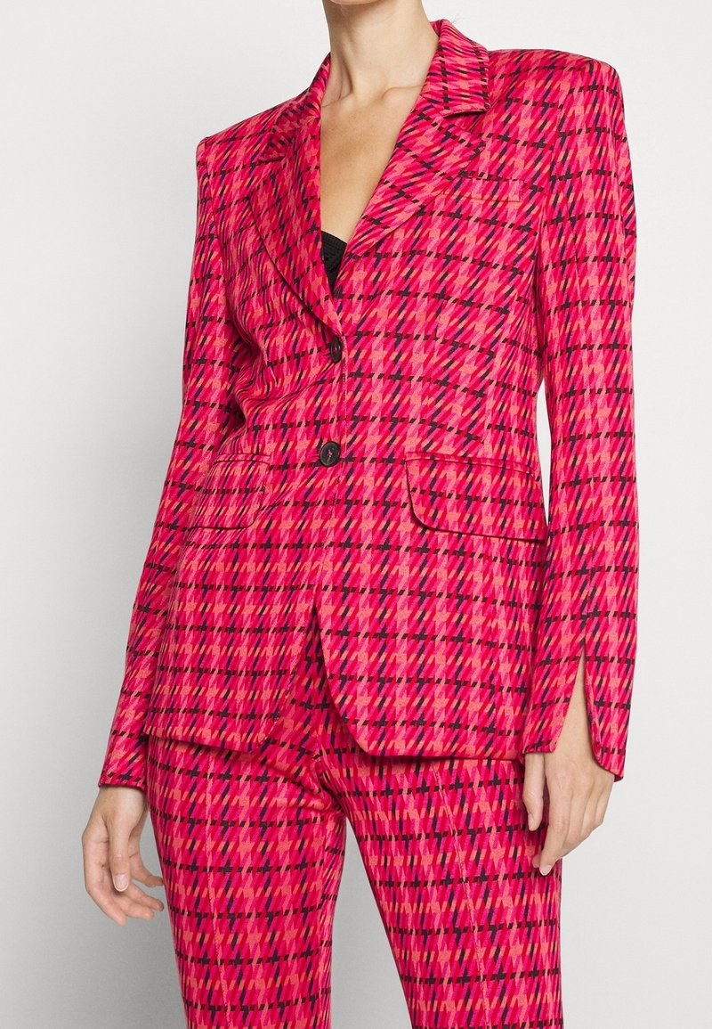 Femme portant un blazer à carreaux rose vif et rouge avec un pantalon assorti, des boutons noirs et un top noir en dessous.